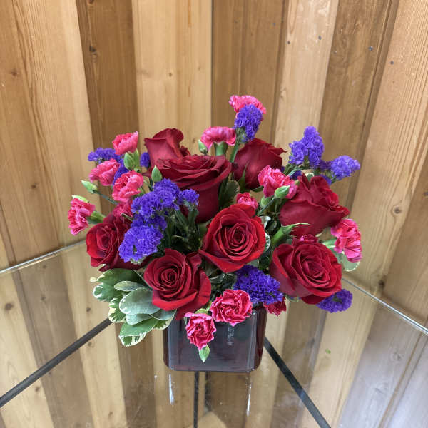 Red roses, pink carnations, and purple flowers arranged in a dark red cube vase