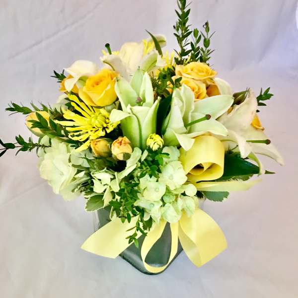 Short arrangement of yellow roses, white lilies, and green hydrangeas in a square glass vase with a yellow ribbon bow