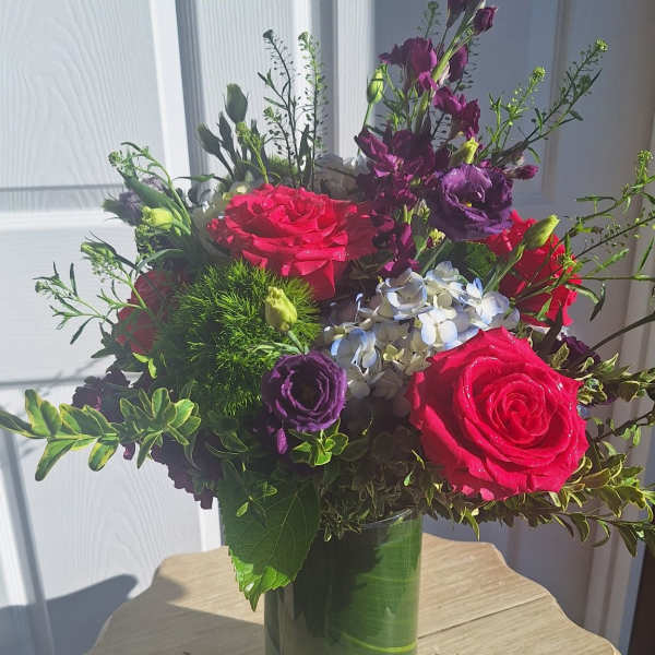 Vibrant arrangement of hot pink roses, purple flowers, and hydrangea in a clear glass vase.
