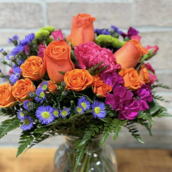 Bright bouquet of orange roses, pink carnations, and purple asters in a clear glass vase