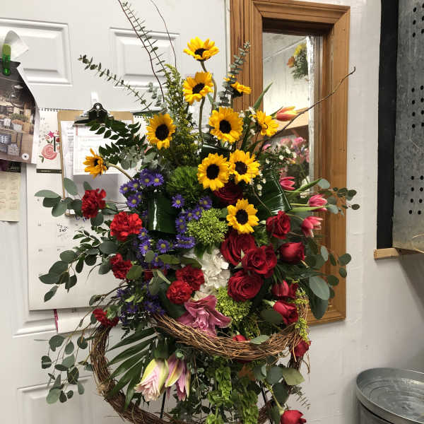 Large standing floral arrangement with sunflowers, red roses, and mixed blooms in a grapevine wreath.