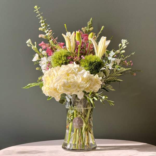 Mixed bouquet with cream hydrangea, lilies, and pink accent flowers in a glass vase