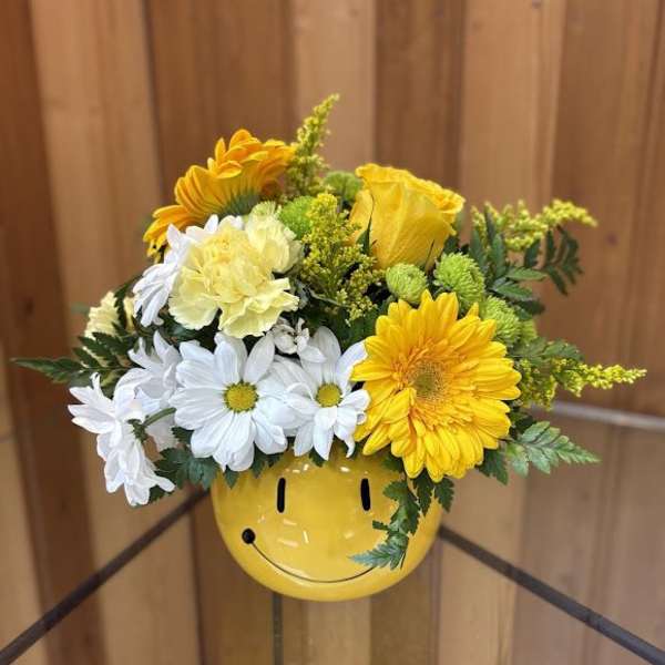Yellow and white daisies, roses, and carnations in a yellow smiley-face ceramic vase.