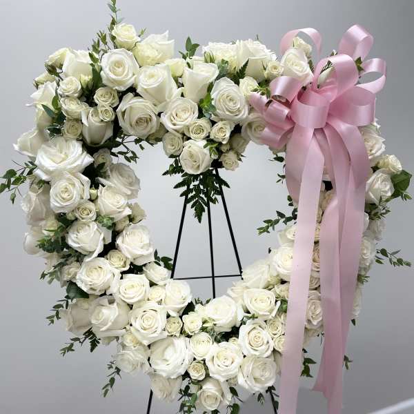 Heart-shaped white rose wreath with a pink ribbon on a stand