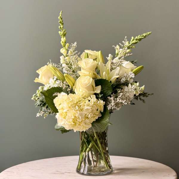 Arrangement of soft yellow roses, lilies, and hydrangeas in a clear glass vase on a round table.