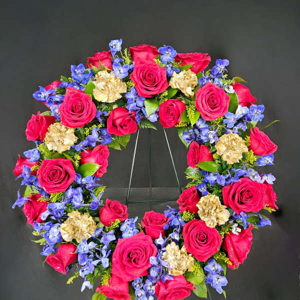 Heart-shaped wreath of pink roses, blue flowers, and gold carnations on a stand