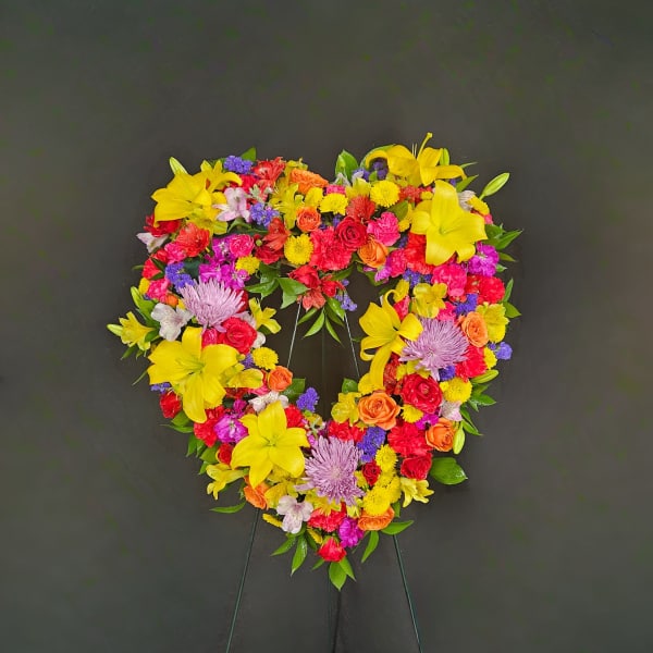 Heart-shaped standing wreath of bright yellow lilies, roses, carnations, and mums on a dark background