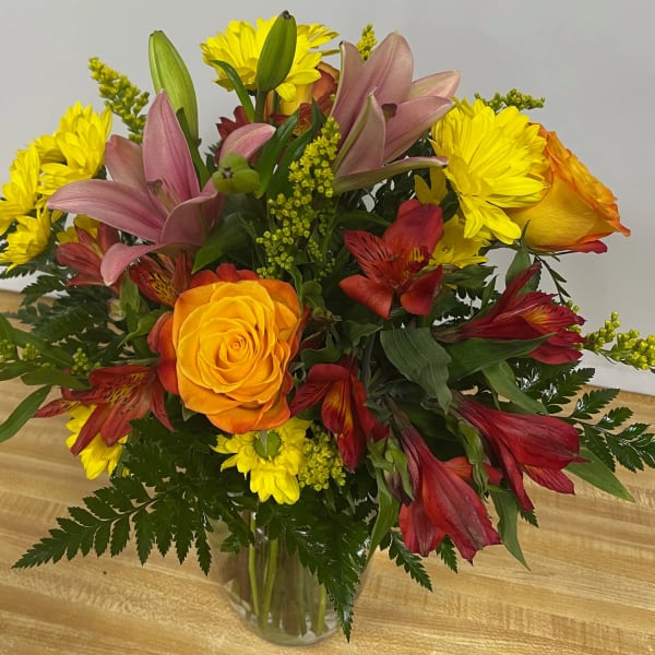 Mixed bouquet of yellow daisies, roses, lilies, and red alstroemeria in a glass vase