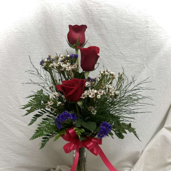 Three red roses in a glass vase with purple filler flowers and a pink ribbon