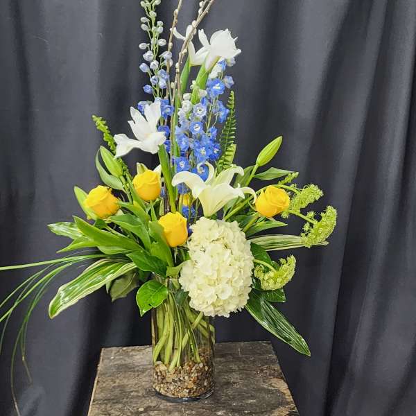 Tall floral arrangement with yellow roses, white lilies, blue flowers, and a white hydrangea in a glass vase