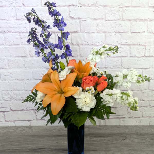 Orange lilies and roses in a tall dark vase with white and purple flowers