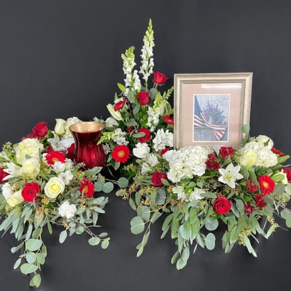 Patriotic red and white floral display with urn and framed flag photo against a black background