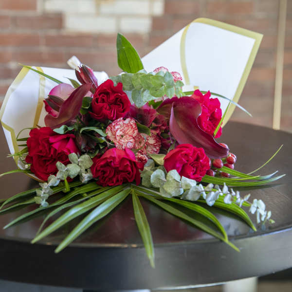Hand-tied bouquet of red roses, burgundy calla lilies, and pink carnations in white wrap.