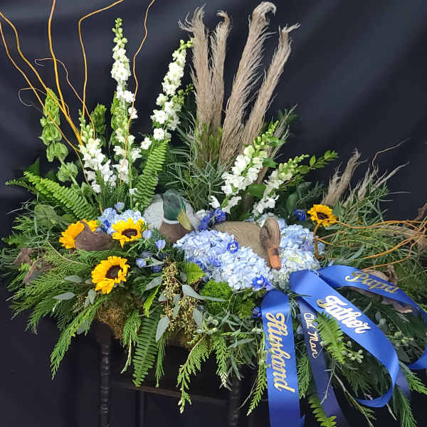 Floral tribute with sunflowers, blue hydrangeas, and blue ribbons on a dark stand