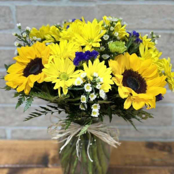 Vase arrangement of bright yellow sunflowers and daisies with white and purple accents.