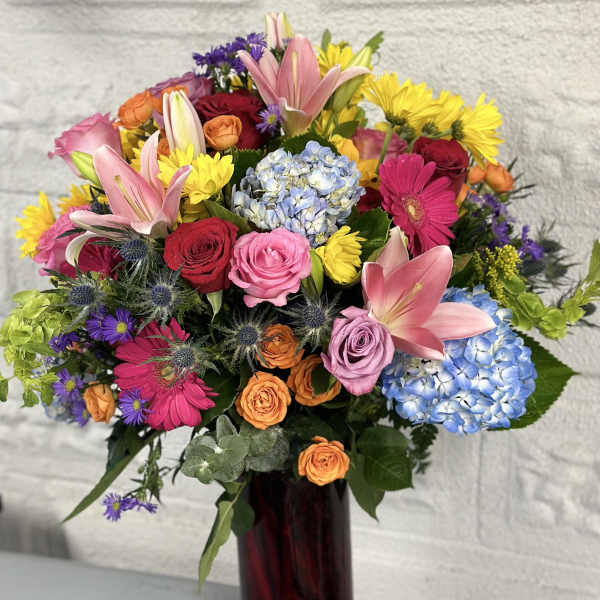 Colorful mixed bouquet in a tall red vase with roses, lilies, hydrangeas, and daisies