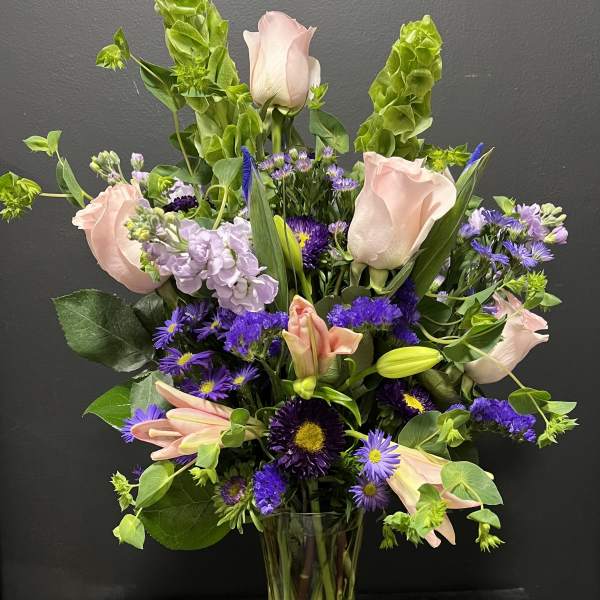 Tall vase arrangement of pink roses and lilies with purple flowers and green spikes.