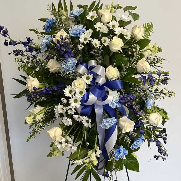 Tall blue and white standing spray with roses, carnations, daisies, and a blue and white bow on an easel.