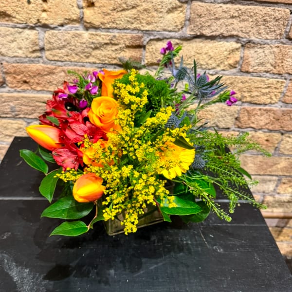 Mixed bouquet of orange roses, tulips, and yellow filler flowers in a glass vase