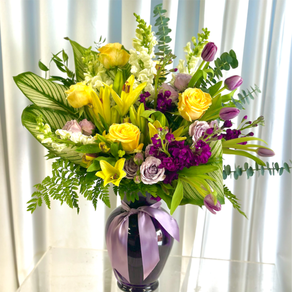 Yellow and purple mixed bouquet in a dark vase with a lavender ribbon