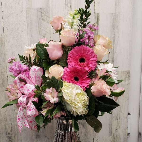 Pink and white mixed flower arrangement in a glass vase with a heart ribbon