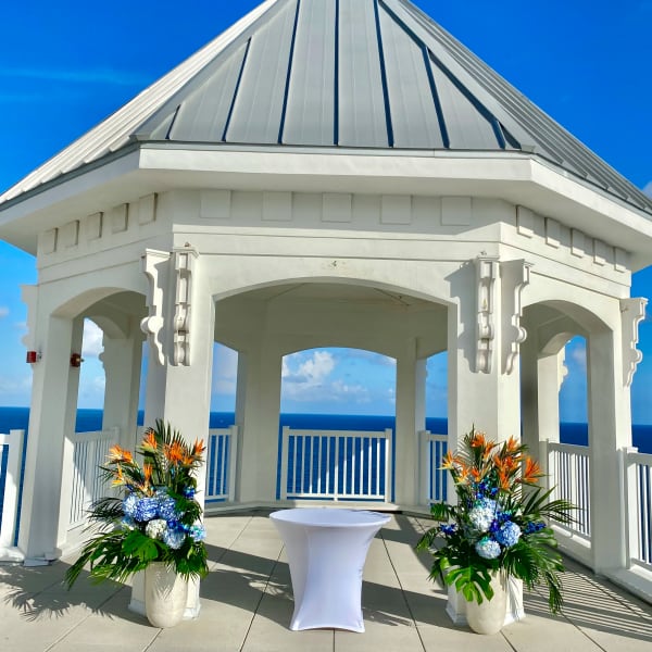Two tropical floral arrangements flank a white cocktail table in a seaside pavilion