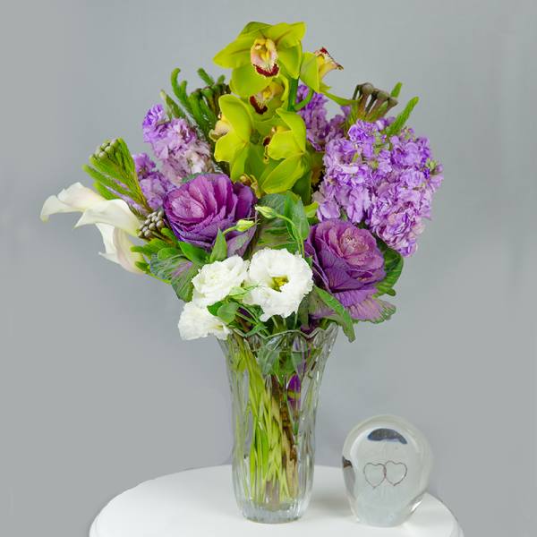Purple and white flower arrangement in a clear glass vase with a small glass keepsake