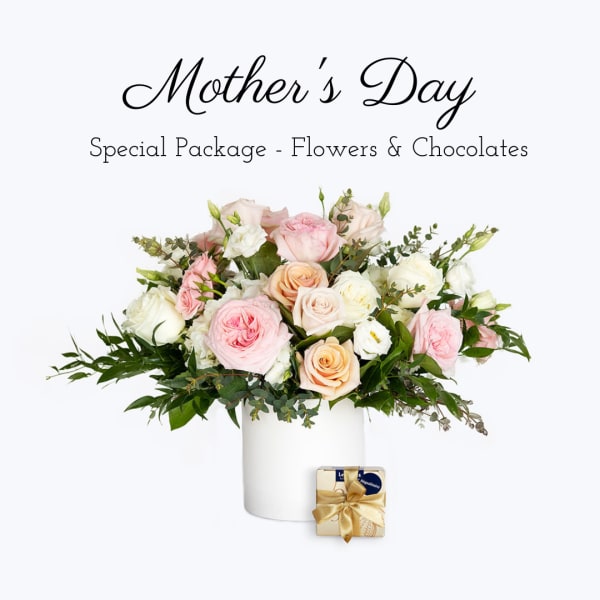 Pastel pink, peach, and white rose arrangement in a white vase with a small gold-wrapped box of chocolates.