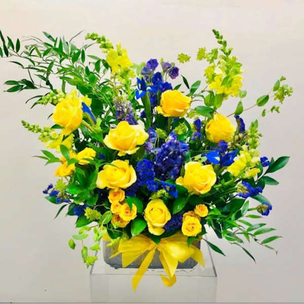 Yellow roses and blue flowers arranged in a square vase with a yellow ribbon.