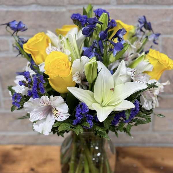 Mixed bouquet of yellow roses, white lilies, and purple flowers in a clear glass vase.