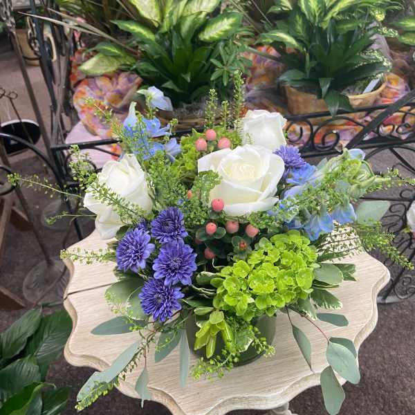 Low arrangement of white roses, purple mums, blue blooms, and green hydrangea in a vase on a small table