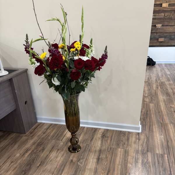 Tall floral arrangement of red roses, yellow blooms, and white flowers in a glass vase
