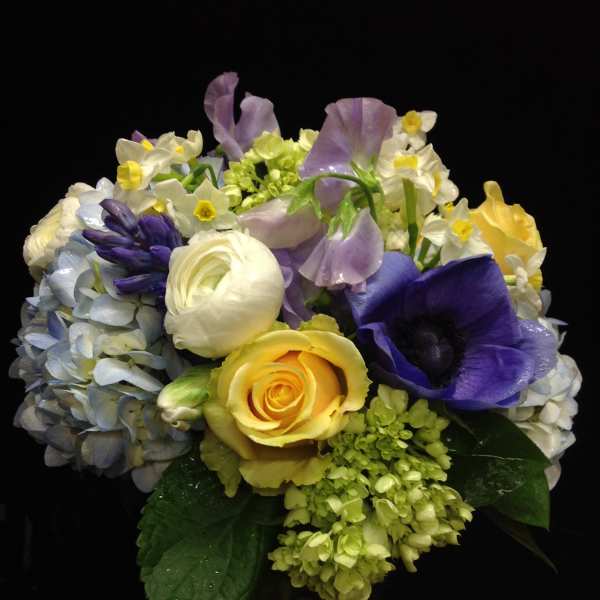 Compact bouquet of yellow roses, white and purple flowers in a glass vase against a dark background