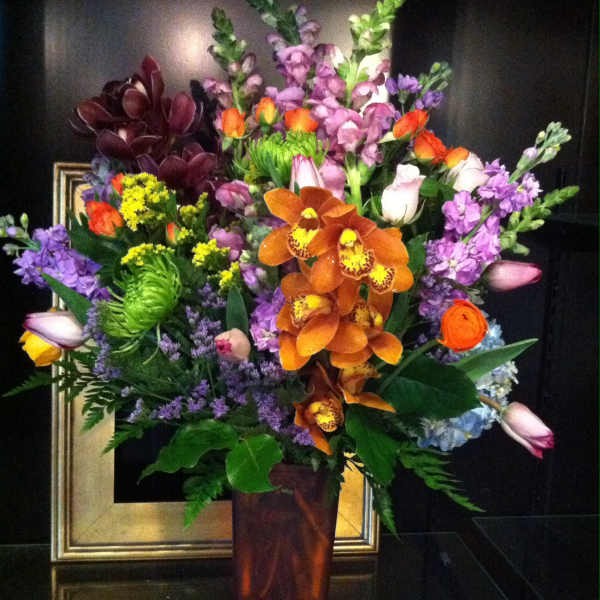 Tall mixed arrangement with orange orchids, purple flowers, and tulips in a brown glass vase