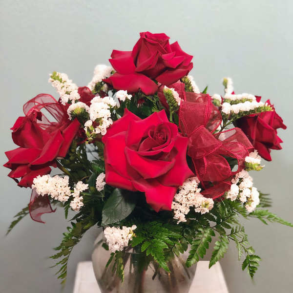 Red roses arranged in a glass vase with white filler flowers and a red ribbon