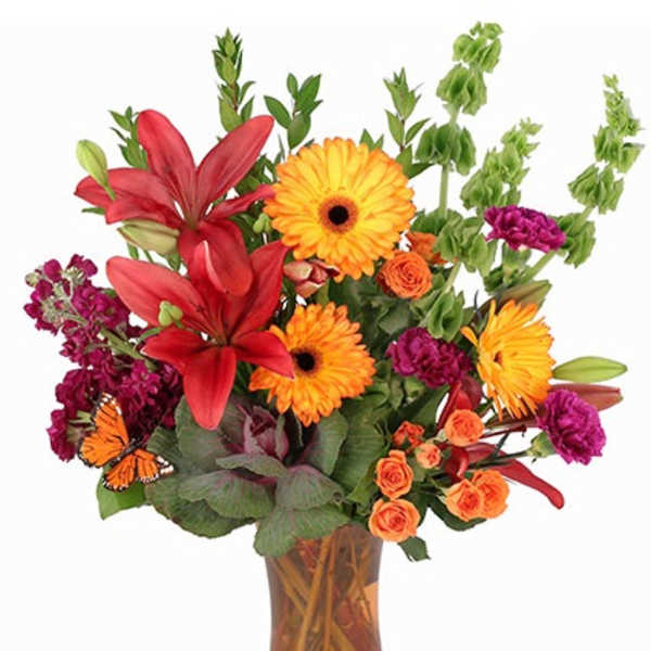 Tall arrangement of orange gerberas, red lilies, and mixed blooms in an amber glass vase.