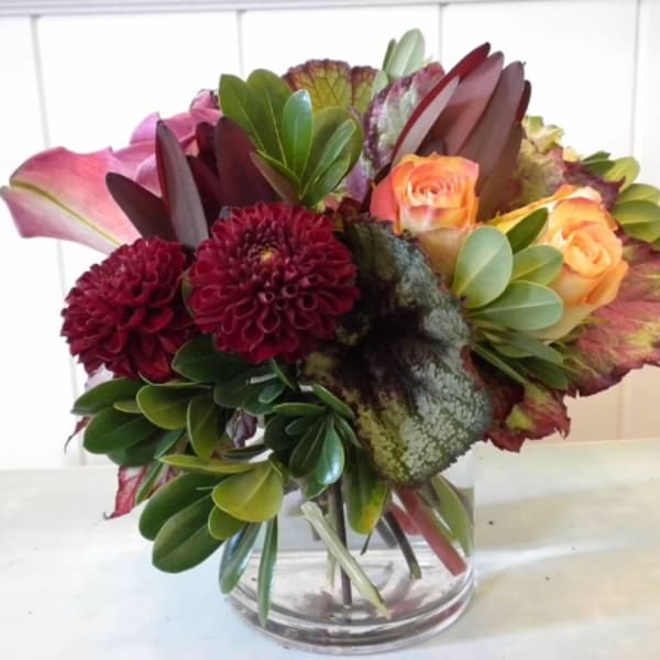 Compact arrangement of burgundy dahlias, peach roses, pink calla lilies, and variegated foliage in a glass vase