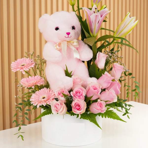 Pink teddy bear surrounded by pink roses and lilies in a white box