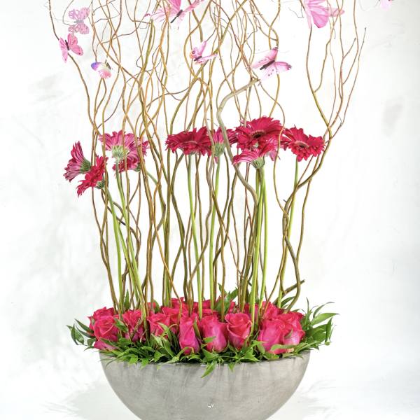 Pink roses and gerbera daisies in a gray bowl with tall curly branches and butterflies