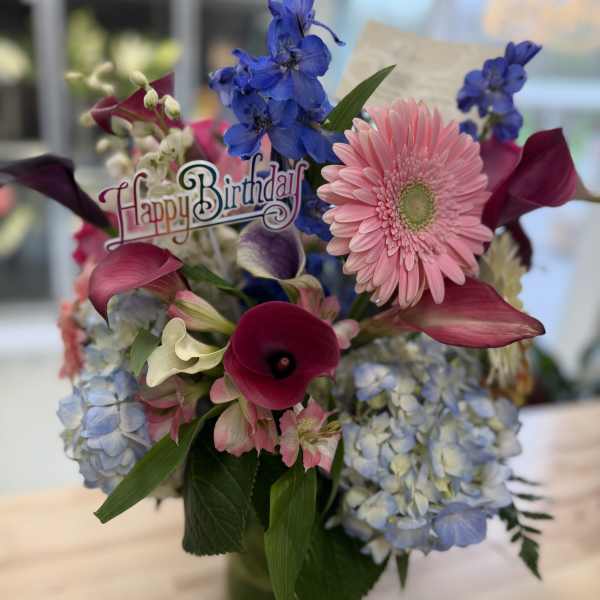 Mixed birthday arrangement with pink gerbera, blue delphinium, hydrangeas, and calla lilies in a clear vase.