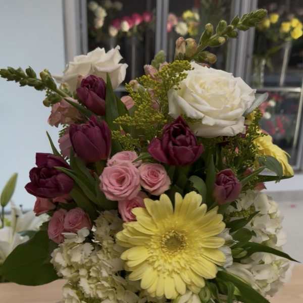 Mixed bouquet of yellow gerbera, white roses, pink spray roses, and dark tulips in a clear glass vase