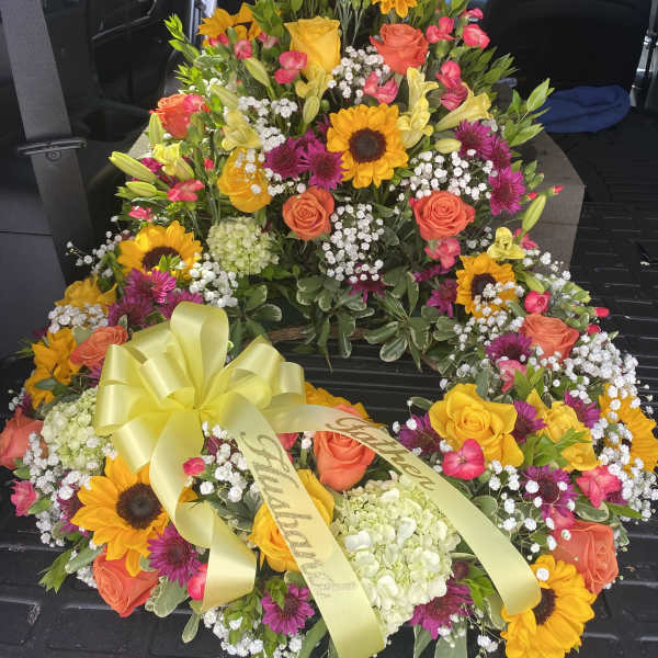 Bright heart-shaped funeral wreath with sunflowers, roses, and a yellow ribbon