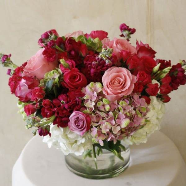 Round arrangement of pink and red roses with hydrangeas and stock in a clear glass cylinder vase