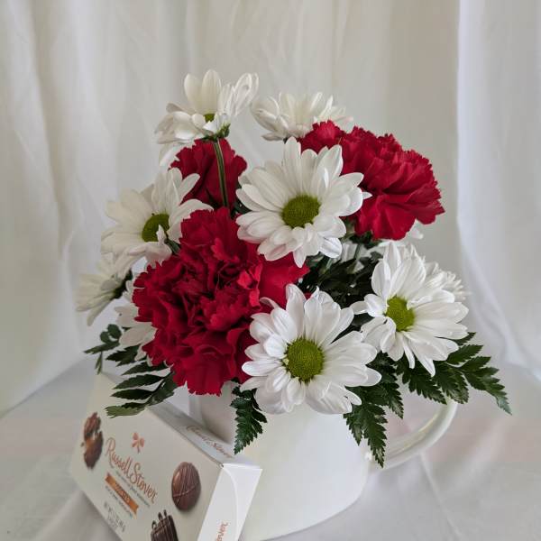 Red carnations and white daisies in a white mug with a chocolate box