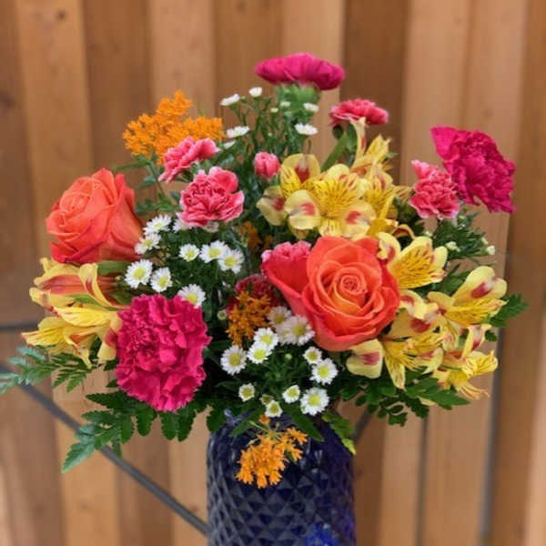 Bright mix of orange roses, pink carnations, yellow alstroemeria, and daisies in a blue glass vase.