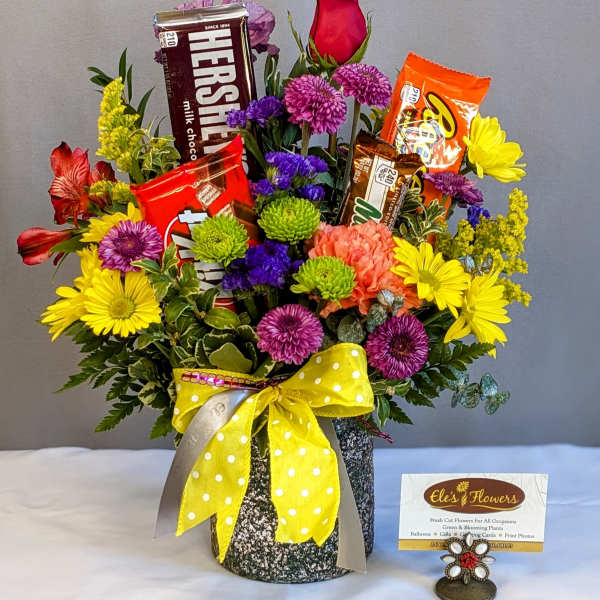 Bright mixed flower arrangement with candy bars in a speckled container tied with a yellow polka-dot bow.