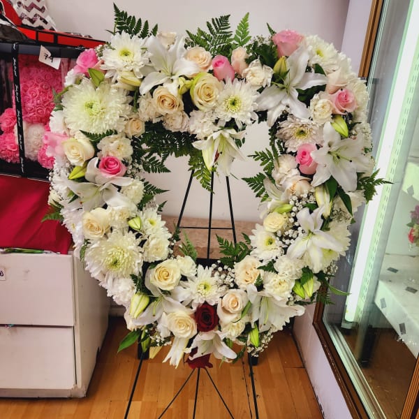 Heart-shaped floral wreath with white lilies, roses, and pink accents on a stand