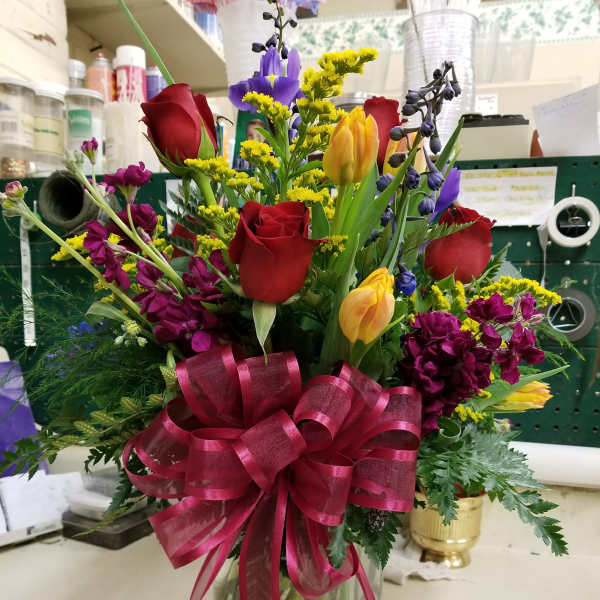 Mixed bouquet of roses, tulips, and purple flowers in a glass vase with a pink ribbon