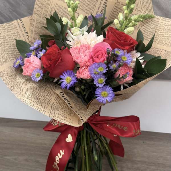Hand-tied bouquet of red roses, pink carnations, and purple asters wrapped in printed paper with a red ribbon