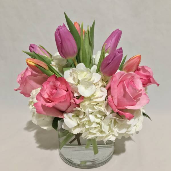 Compact arrangement of pink roses, purple tulips, and white hydrangeas in a clear glass vase